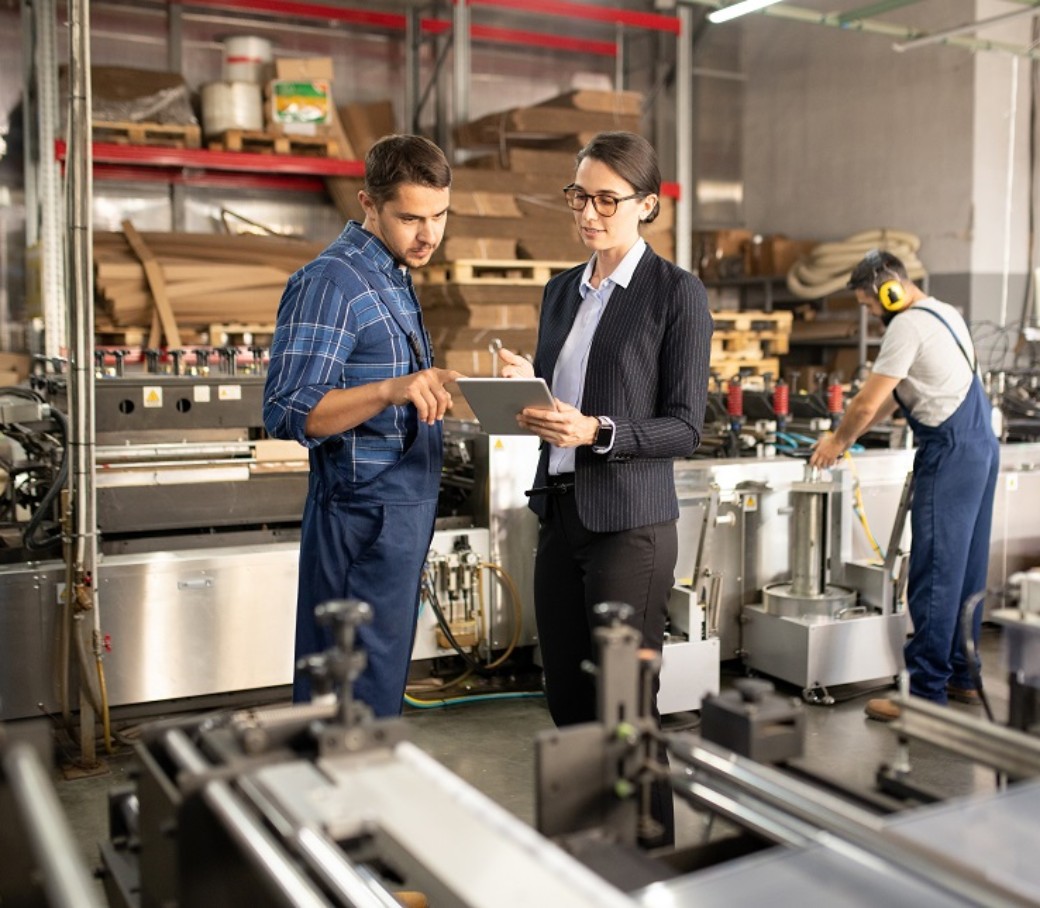Eine Business-Frau und ein Bauarbeiter sprechen zusammen über ein Dokument, welches die Frau in der Hand hält. Die Umgebung ist eine Fabrik. 