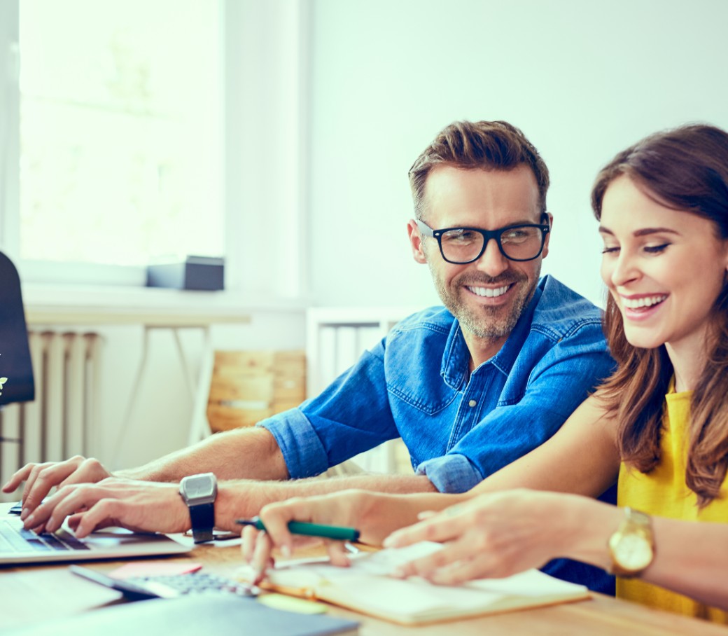 Ein Mann und eine Frau sitzen nebeneinander und unterhalten sich lachend, während sie an Unterlagen und am Laptop arbeiten. 