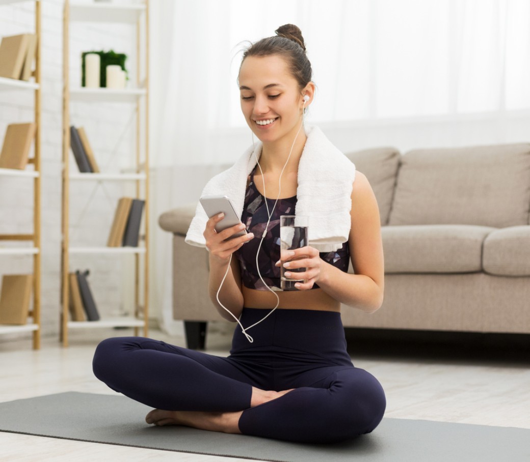 Eine Frau sitzt im Schneidersitz auf einer Sportmatte und lacht in ihr Handy.