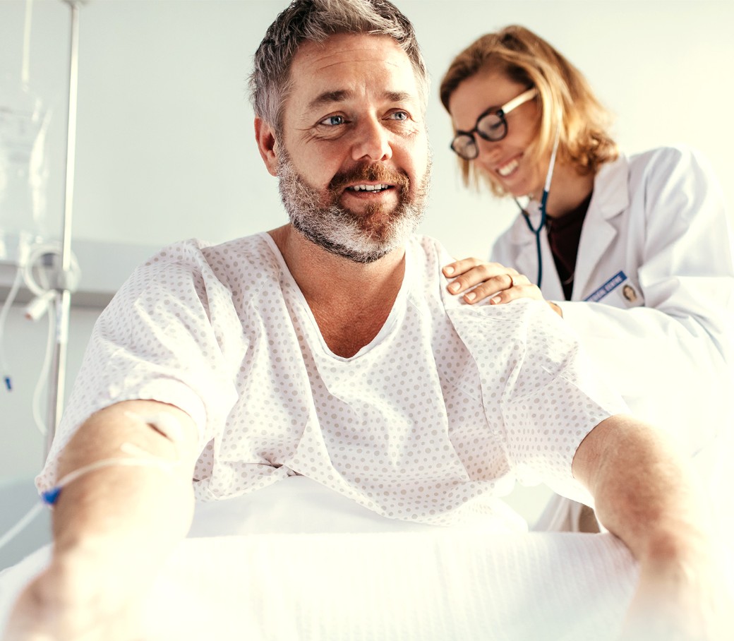 Eine Ärztin hört mit einem Stethoskop den Rücken eines männlichen Patienten ab, welcher aufrecht im Patientenbett sitzt.