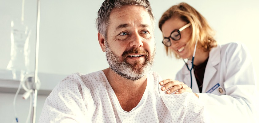 Eine Ärztin hört mit einem Stethoskop den Rücken eines männlichen Patienten ab, welcher aufrecht im Patientenbett sitzt.